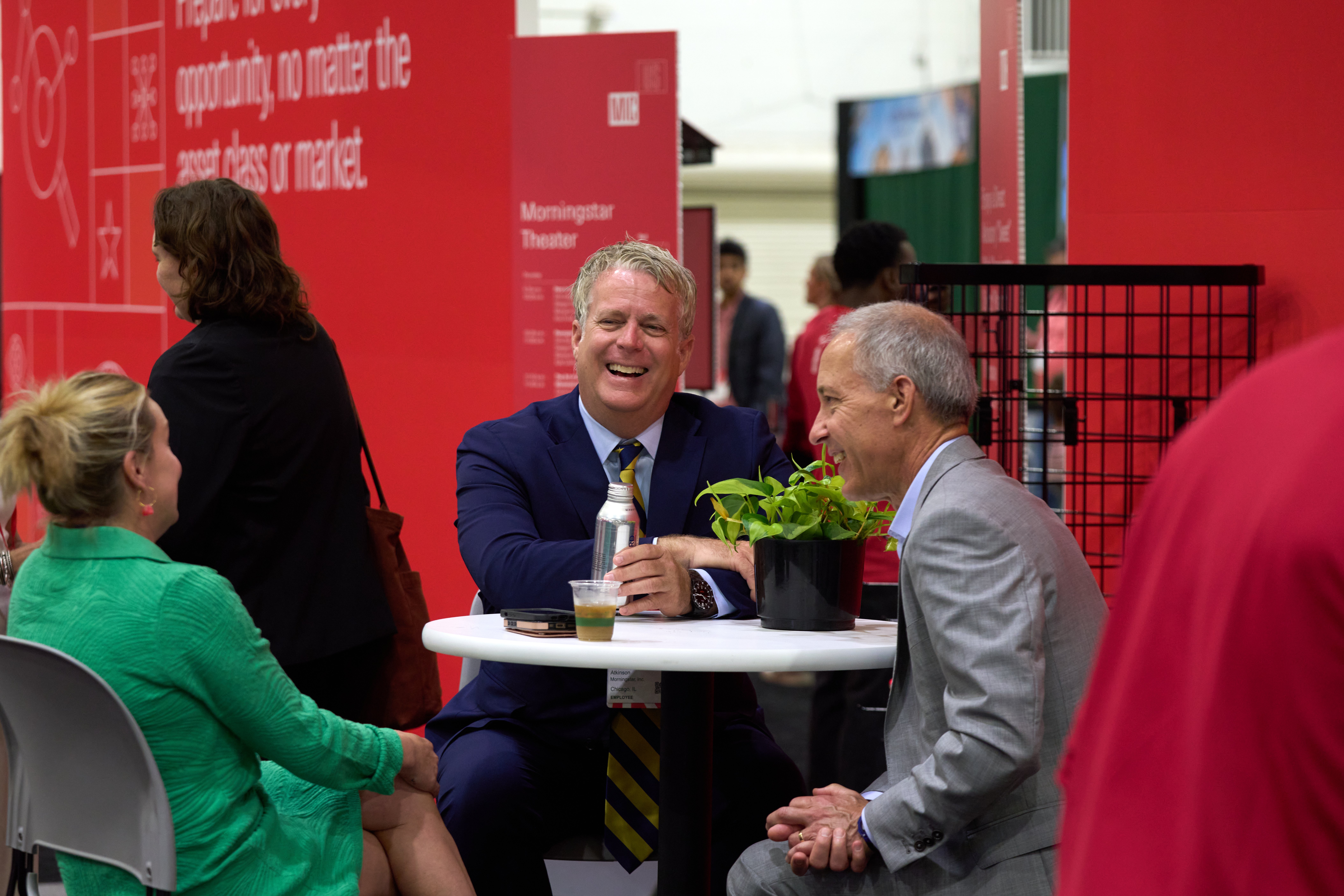 Networking. A group of professional attendees engage in a cheerful conversation around a small white bistro table at a Morningstar Investment conference. A man in a blue suit and gold-and-blue striped tie laughs heartily while holding a silver water bottle, seated across from a man in a grey suit and next to a woman in a bright green textured dress. The setting features vibrant red backdrop panels with white text and a small potted plant on the table, capturing a lively networking moment in a busy event space.