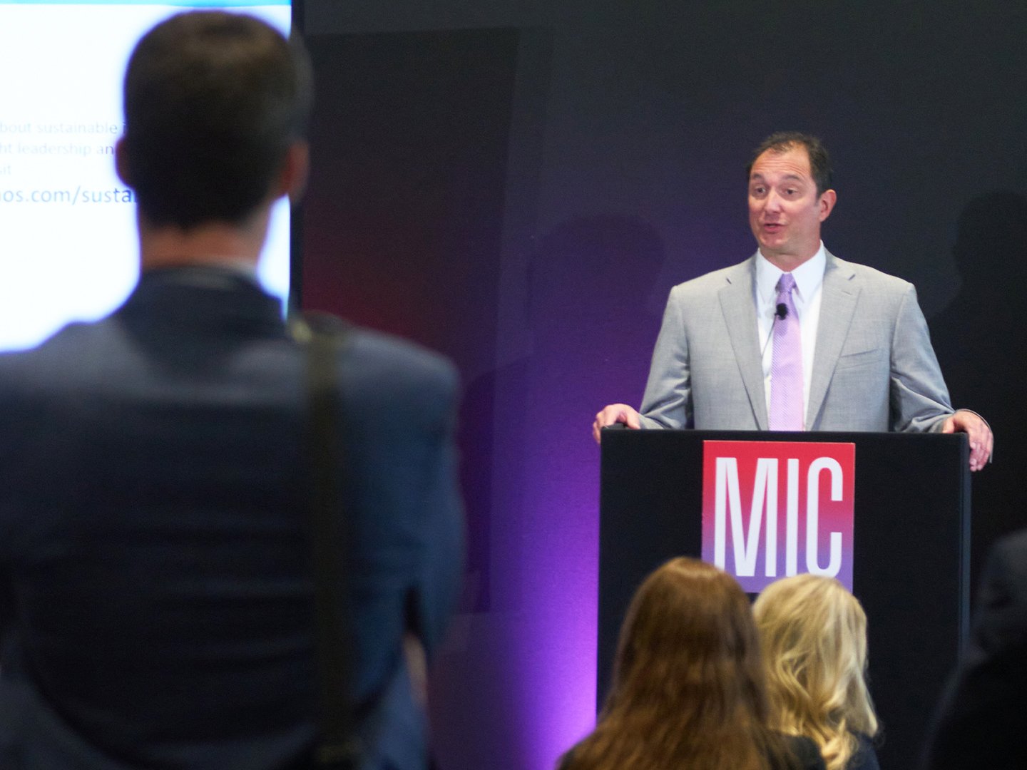 A speaker presenting from a podium with an MIC logo while attendees look on.