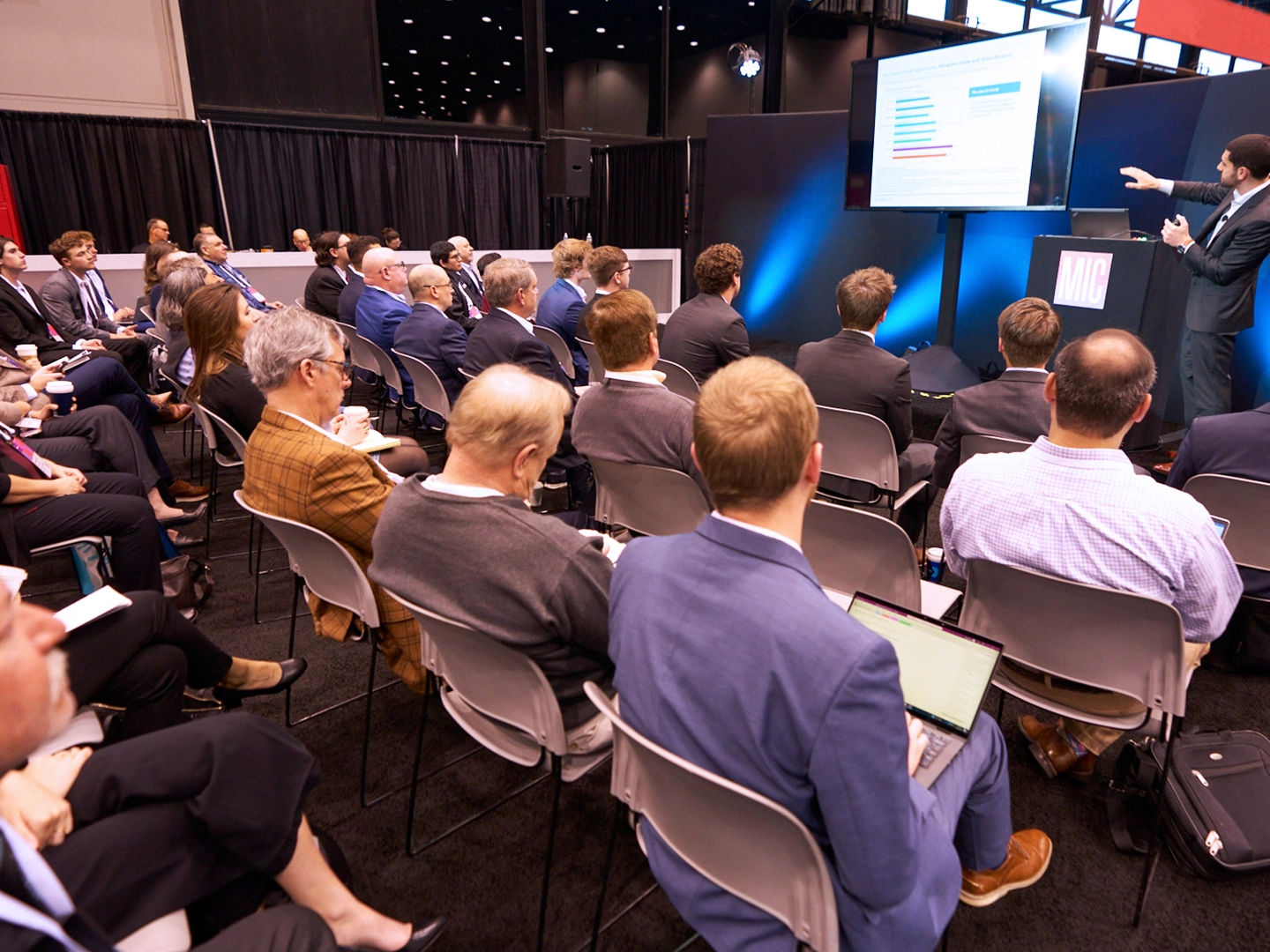 Audience watching a presenter at a sponsor-branded stage with projection screens